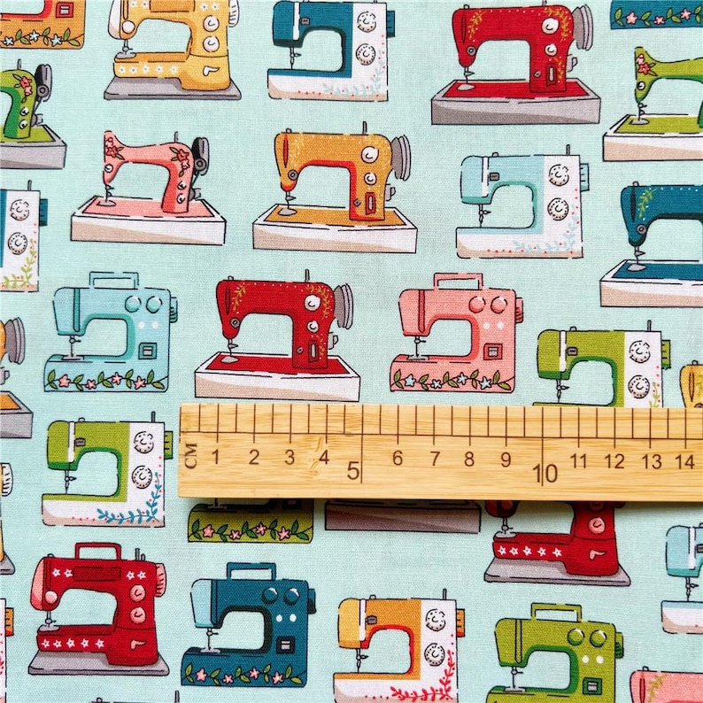 May include: A turquoise fabric with a repeating pattern of colorful vintage sewing machines. The fabric is measured with a wooden ruler with markings in centimeters.