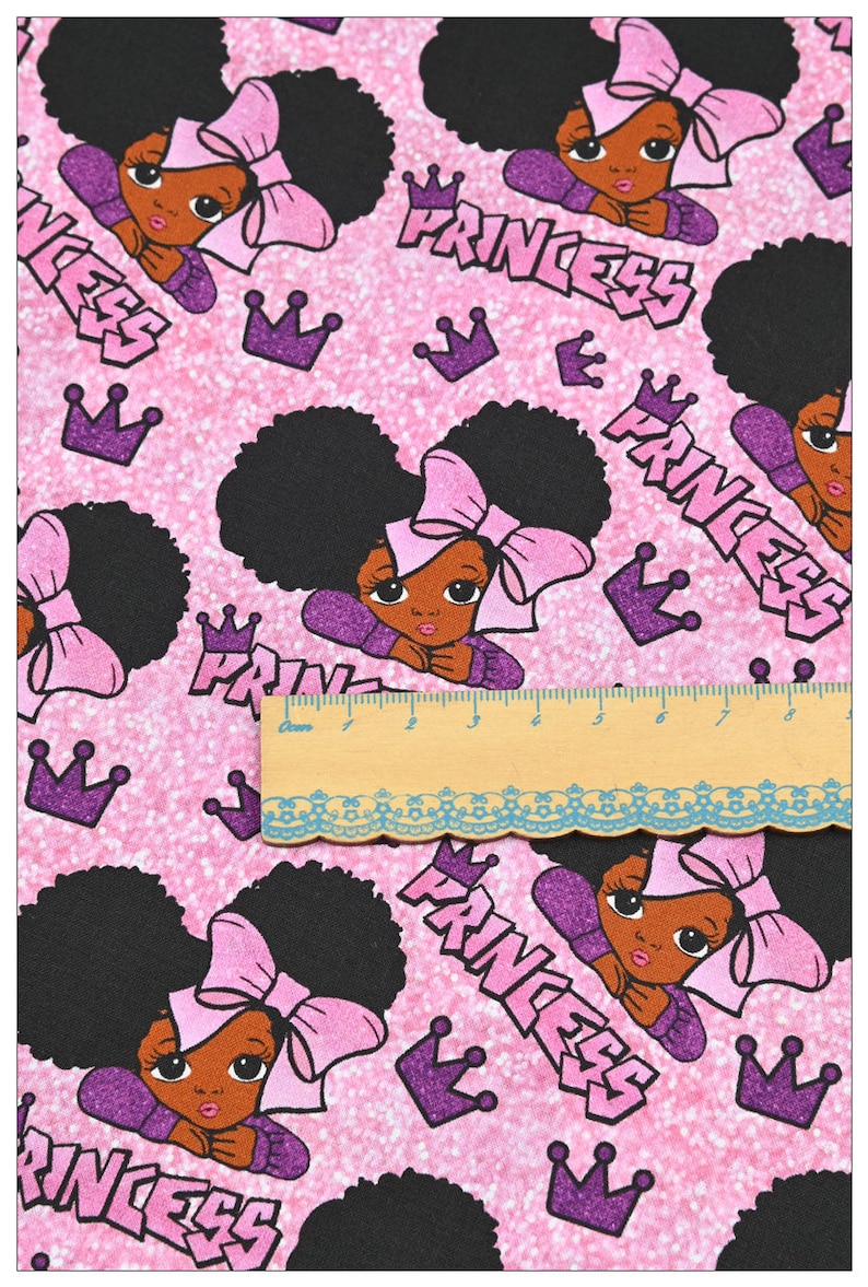 May include: Pink fabric with a repeating pattern of black girls with pink bows and the word "Princess" in purple letters. The background is a sparkly pink.
