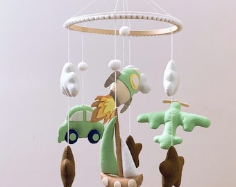 Puede incluir: Móvil para bebé con recortes de fieltro de un coche verde, un cohete, un avión, un velero, nubes y estrellas. El móvil está suspendido de un anillo de madera con cuentas blancas.
