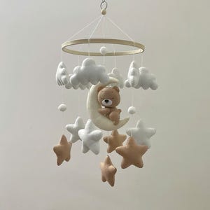 May include: A baby mobile featuring a wooden ring with hanging felt clouds, stars, and a crescent moon. A small brown teddy bear sits on the moon. The stars are in white and tan colors. The mobile is suspended from a string.