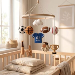 May include: A sports-themed nursery mobile featuring felt hanging ornaments including a soccer ball, basketball, football, baseball, a blue jersey with 01 MVP text, a baseball bat, a foam finger with We're #1 text, a glove, a trophy, and a white cloud.