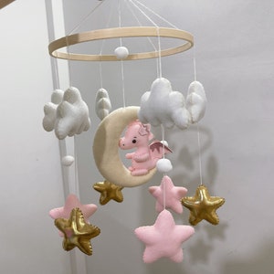 May include: A pink and white baby mobile with a crescent moon, stars, and clouds. The moon has a pink dragon on it. The stars are gold and the clouds are white.
