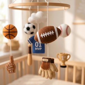 May include: A sports-themed nursery mobile featuring a circular wooden frame with hanging felt ornaments including a basketball, soccer ball, football, baseball, a blue jersey with 01 MVP text, a baseball bat, a glove, a trophy, and a foam finger.