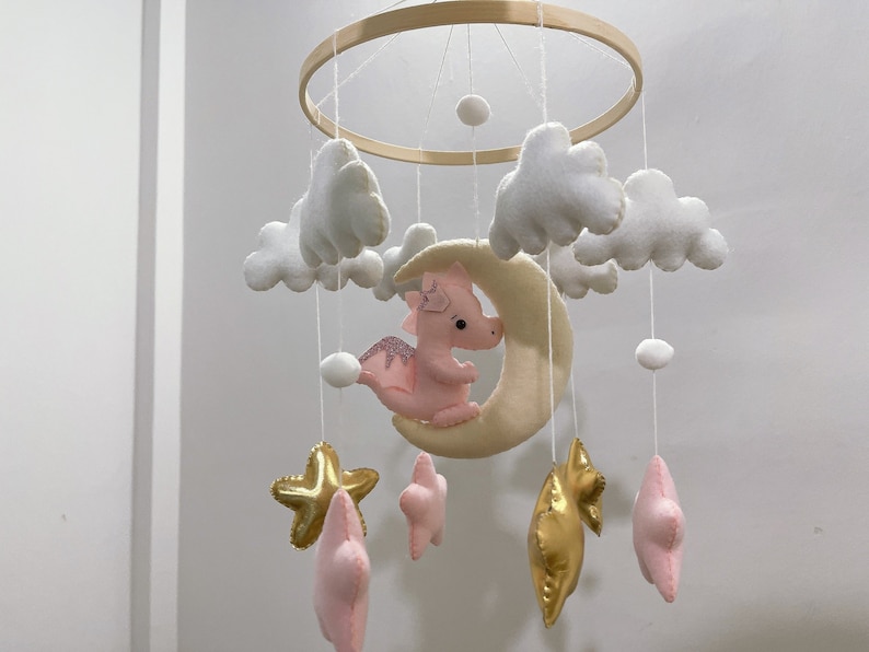 May include: A pink and white baby mobile with a crescent moon, stars, and clouds. The moon has a pink dragon on it.