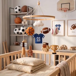 May include: A sports-themed nursery mobile featuring felt hanging ornaments including a soccer ball, basketball, football, baseball, a blue jersey with '01 MVP' text, a brown baseball bat, a 'We're #1' foam finger, a glove, a trophy, and a white cloud.