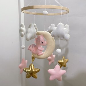 May include: A pink felt dragon with gold glitter wings hangs from a wooden mobile with white felt clouds and gold star shapes.