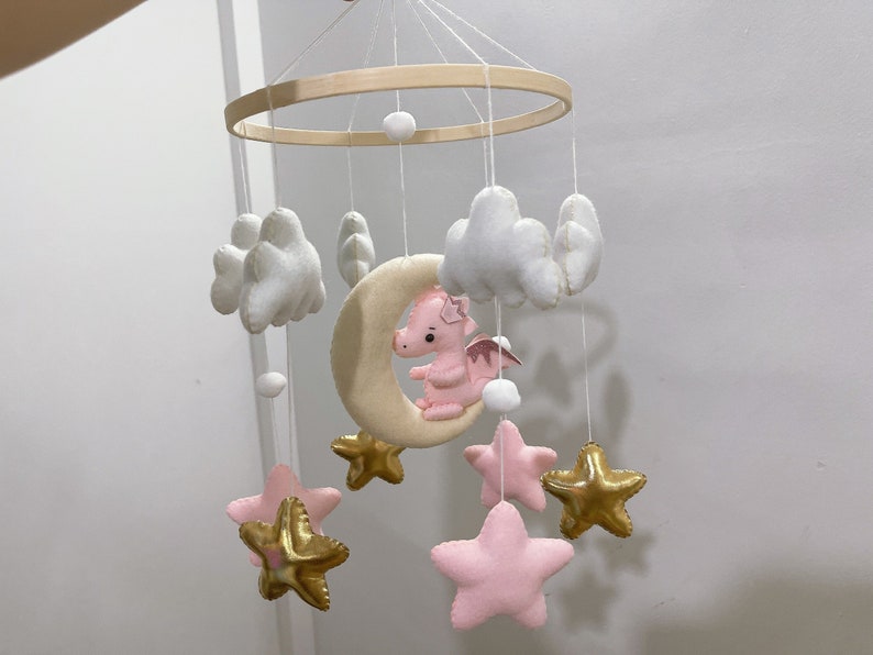 May include: A pink and white baby mobile with a crescent moon, stars, and clouds. The moon has a pink dragon on it. The stars are gold and pink.