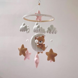 May include: A white felt baby mobile with a brown bear sleeping on a white crescent moon. The mobile also features pink and brown stars, white clouds, and white pom-poms.