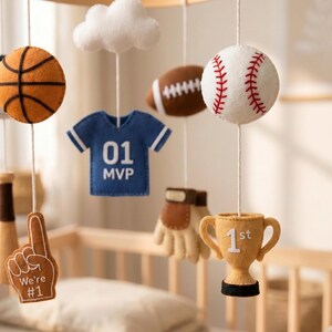 May include: A hanging nursery mobile featuring felt sports-themed elements including a basketball, a white baseball with red stitching, a brown football, a blue jersey with 01 MVP, a tan trophy with 1st, a foam finger with We're #1, and a white cloud.