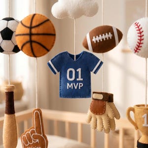 May include: A hanging nursery mobile featuring felt sports-themed ornaments including a soccer ball, basketball, football, baseball, a blue jersey with the text 01 MVP, a baseball bat, a foam finger, a baseball glove, a trophy, and a white cloud.