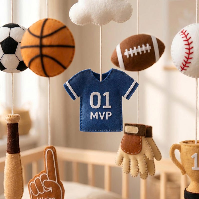 May include: A hanging nursery mobile featuring felt sports-themed ornaments including a soccer ball, basketball, football, baseball, a blue jersey with the text 01 MVP, a baseball bat, a foam finger, a baseball glove, a trophy, and a white cloud.
