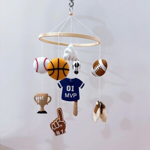 May include: A sports-themed hanging mobile featuring a circular wooden frame with felt ornaments including a baseball, basketball, soccer ball, football, a blue jersey with 01 MVP text, a tan trophy with 1st text, a foam finger, and a glove.