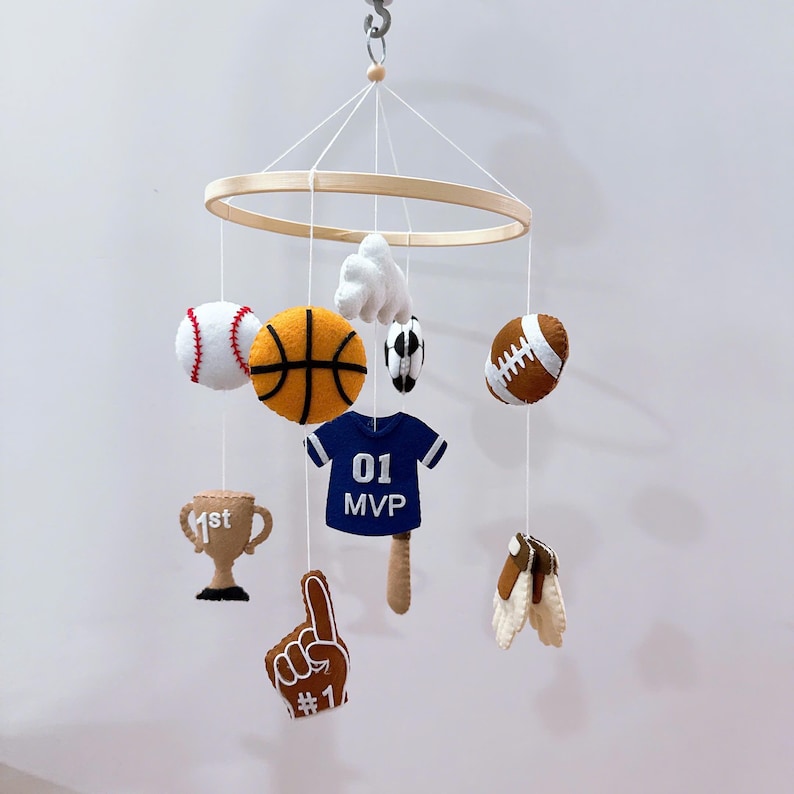 May include: A sports-themed hanging mobile featuring a circular wooden frame with felt ornaments including a baseball, basketball, soccer ball, football, a blue jersey with 01 MVP text, a tan trophy with 1st text, a foam finger, and a glove.
