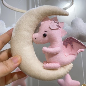 May include: A pink felt dragon with glitter wings is hanging from a white felt crescent moon mobile. The mobile also includes white felt clouds.