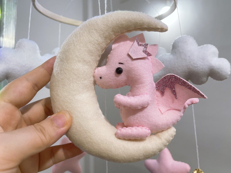 May include: A pink felt dragon with glitter wings is hanging from a white felt crescent moon mobile. The mobile also includes white felt clouds.