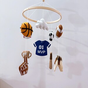 May include: A sports-themed nursery mobile featuring a circular wooden frame with hanging felt ornaments including a basketball, a white cloud, a football, a soccer ball, a blue jersey with 01 MVP text, a baseball bat, a trophy, and a foam finger.