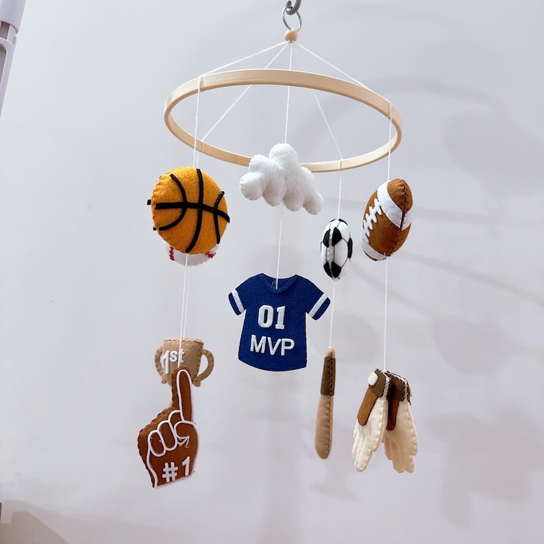 May include: A hanging mobile featuring felt sports-themed ornaments suspended from a circular wooden frame. The items include a basketball, a white cloud, a football, a soccer ball, a blue jersey with 01 MVP text, a trophy, a foam finger with #1, a bat, and gloves.