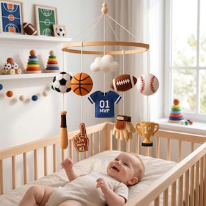 May include: A nursery mobile featuring felt sports-themed ornaments including a soccer ball, basketball, football, baseball, a blue jersey with '01 MVP' text, a baseball bat, a 'We're #1' foam finger, a baseball glove, a trophy, and a white cloud.