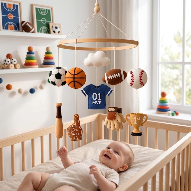 May include: A nursery mobile featuring felt sports-themed ornaments including a soccer ball, basketball, football, baseball, a blue jersey with '01 MVP' text, a baseball bat, a 'We're #1' foam finger, a baseball glove, a trophy, and a white cloud.