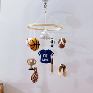 May include: A sports-themed hanging mobile featuring felt shapes suspended from a circular wooden frame. The items include a basketball, baseball, football, soccer ball, a trophy, a foam finger, a baseball bat, gloves, and a blue jersey with 01 MVP.