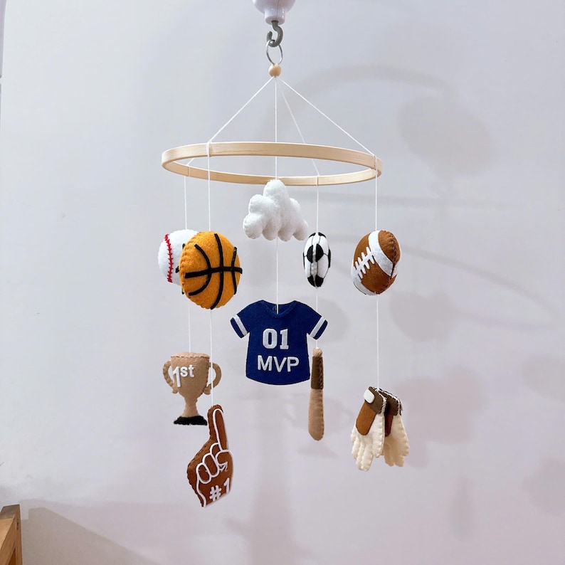 May include: A sports-themed hanging mobile featuring felt shapes suspended from a circular wooden frame. The items include a basketball, baseball, football, soccer ball, a trophy, a foam finger, a baseball bat, gloves, and a blue jersey with 01 MVP.