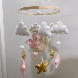May include: A white and pink baby mobile with a crescent moon, clouds, stars, and a pink dragon. The mobile is made of felt and has a wooden ring.