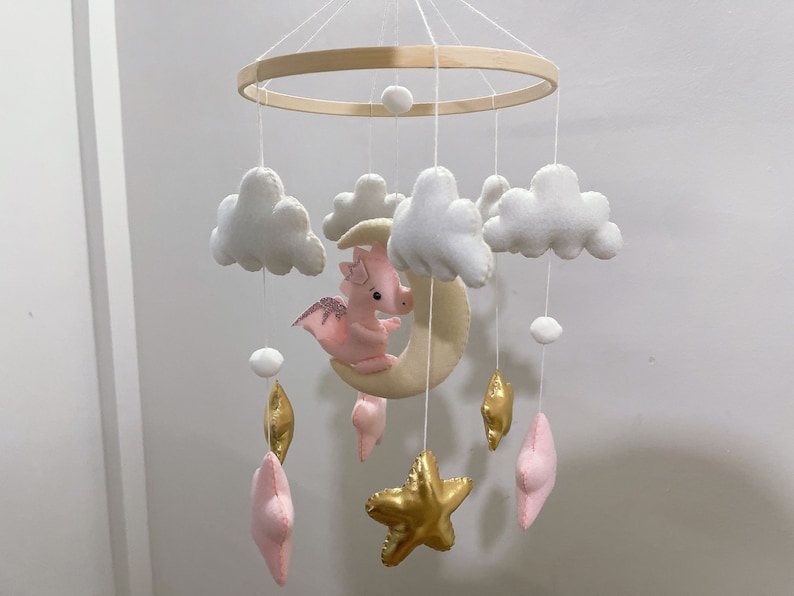 May include: A white and pink baby mobile with a crescent moon, clouds, stars, and a pink dragon. The mobile is made of felt and has a wooden ring.