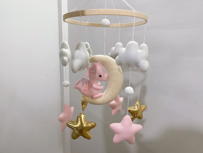 May include: A pink felt dragon with gold glitter wings hangs from a wooden mobile with white felt clouds and gold star shapes.