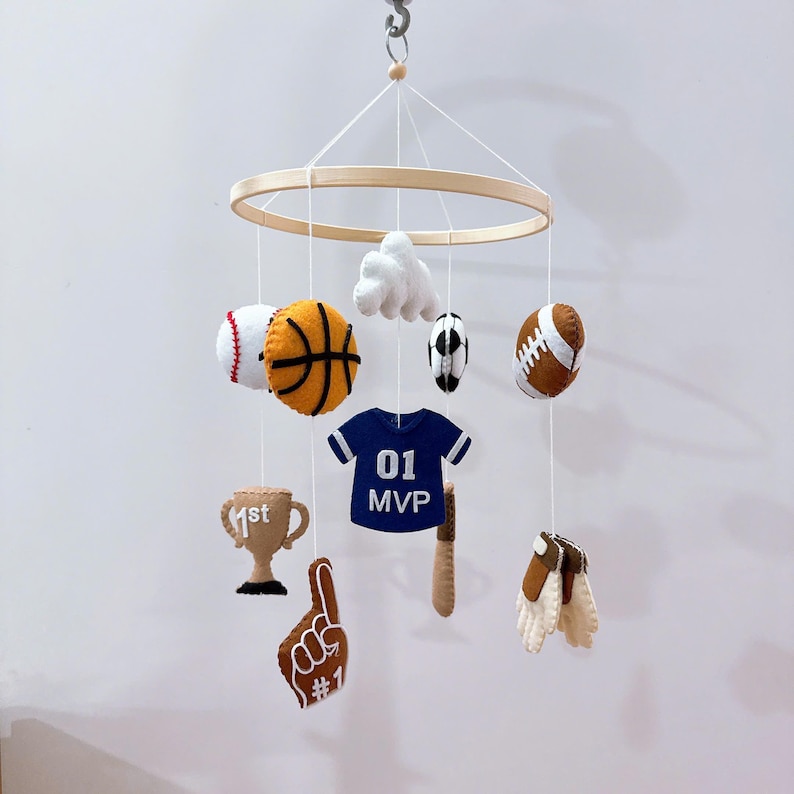 May include: A sports-themed hanging mobile featuring felt shapes suspended from a circular wooden frame. The items include a baseball, basketball, soccer ball, football, a blue jersey with 01 MVP text, a trophy with 1st text, a foam finger, and gloves.