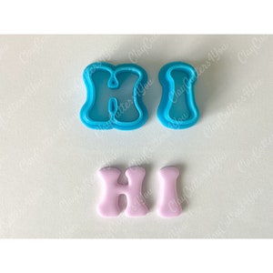 May include: Two blue plastic cookie cutters shaped like the letters "H" and "I" and two pink fondant letters "H" and "I" on a white background.