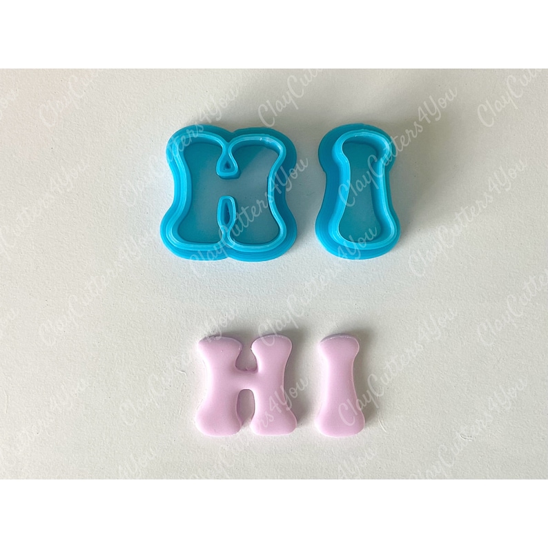 May include: Two blue plastic cookie cutters shaped like the letters "H" and "I" and two pink fondant letters "H" and "I" on a white background.