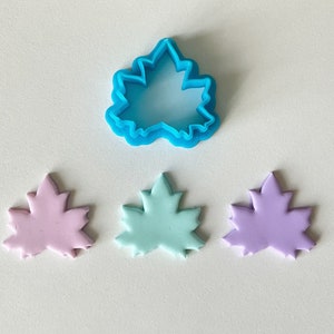 May include: Three maple leaf shapes made from polymer clay in pink, light blue, and purple. A blue plastic maple leaf cookie cutter is above the clay shapes.