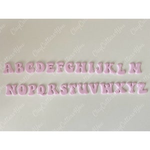 May include: Pink alphabet letter cookie cutters. The letters are in a stylized font and are arranged in two rows. The letters are A through Z.