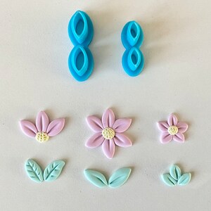 Può includere: Due tagliapasta per fiori in plastica blu e sei fiori e foglie di pasta di zucchero. I fiori sono rosa e le foglie sono blu chiaro.