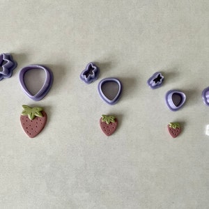 Könnte beinhalten: Ein Set aus violetten Plastik-Ausstechern in Form eines Schmetterlings, einer Erdbeere, einer Blume und Blättern. Außerdem gibt es drei kleine, braune, tonartige Erdbeeren.