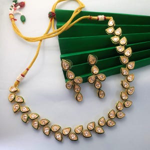 Op de afbeelding: Een goudkleurige ketting- en oorbellenset. De sieraden hebben druppelvormige, heldere edelstenen met een gouden rand. De ketting en oorbellen worden gepresenteerd op een groene achtergrond. De ketting heeft een gouden koord.