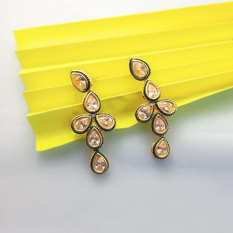Op de afbeelding: Een paar goudkleurige oorhangers met een bloemmotief. Elke oorbel heeft meerdere peervormige, heldere edelstenen gezet in een gouden frame met een donkergroene rand. De oorbellen worden gepresenteerd op een gele achtergrond.
