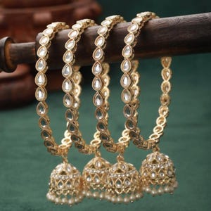 Puede incluir: Conjunto de cuatro brazaletes dorados con piedras blancas en forma de pera y campanillas colgantes con detalles de perlas.