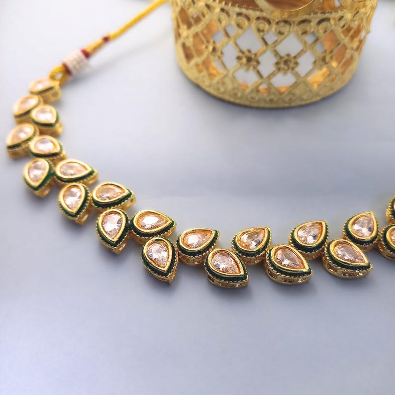 Op de afbeelding: Een goudkleurige ketting met druppelvormige hangers. Elke hanger heeft een licht perzikkleurige steen, gezet in een gouden frame met een groene rand. De ketting ligt op een lichtblauwe ondergrond, met een decoratieve gouden container op de achtergrond.