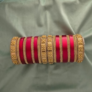 May include: A collection of red and gold bangles. The bangles feature a striking red central band, complemented by ornate gold detailing. The gold sections are embellished with tiny, shimmering accents, giving a luxurious feel. The bangles are presented on a soft, green fabric.
