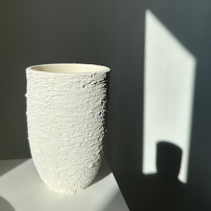 May include: White ceramic vase with a textured surface. The vase is tall and cylindrical with a slightly rounded bottom.