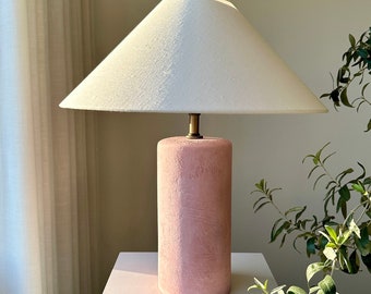 May include: A pink ceramic table lamp with a white linen lampshade. The lamp is on a white pedestal.