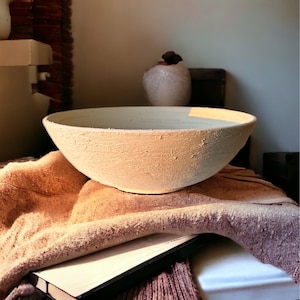 May include: A large, off-white ceramic bowl with a textured surface sits on a brown and beige blanket. A black book is partially visible under the blanket.