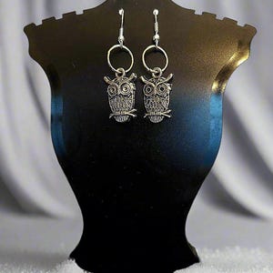 May include: Silver owl earrings with a circular hoop design. The owls are detailed with textured feathers and large eyes.