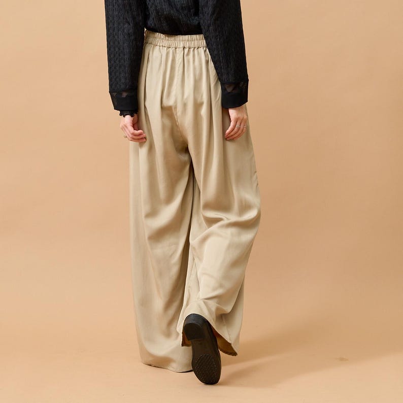 Puede incluir: Pantalones anchos color beige con cintura el&aacute;stica. Los pantalones est&aacute;n hechos de una tela fluida y se combinan con una parte superior negra de manga larga. El modelo lleva zapatos negros. El conjunto se presenta sobre un fondo neutro.