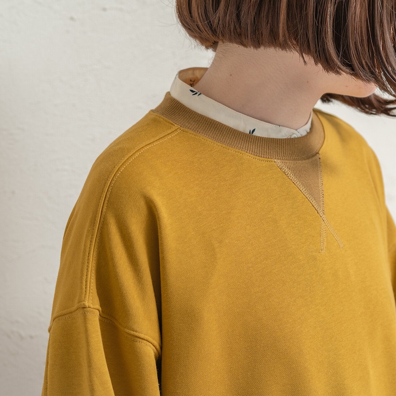 May include: A mustard yellow sweatshirt with a brown ribbed crew neck and a brown triangle detail on the neckline.
