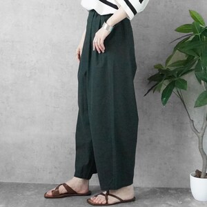 May include: Dark green wide-leg pants paired with brown leather sandals. The pants have a high waist and a loose fit. A black and white striped top is partially visible. A silver bracelet and rings are worn. A potted plant is in the background.