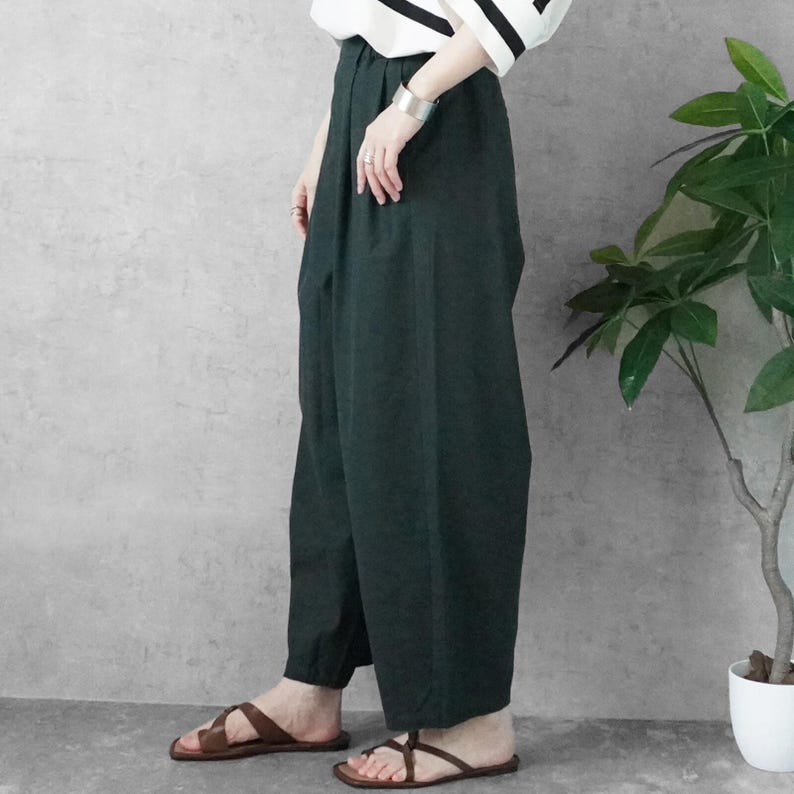 May include: Dark green wide-leg pants paired with brown leather sandals. The pants have a high waist and a loose fit. A black and white striped top is partially visible. A silver bracelet and rings are worn. A potted plant is in the background.
