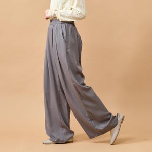 Puede incluir: Pantalones grises de pernera ancha con cintura alta y bolsillos. Los pantalones est&aacute;n hechos de una tela fluida y se combinan con una parte superior color crema y zapatos de color claro. Los pantalones tienen un corte relajado.