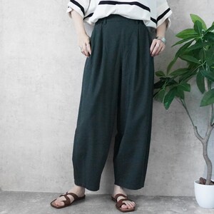 May include: Dark green wide-leg pants with a high waist. The pants are paired with a white and black striped short-sleeve shirt and brown sandals. A plant in a white pot is in the background.